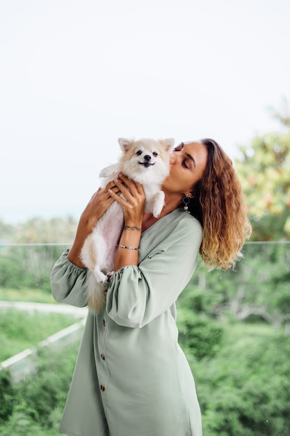 Retrato ao ar livre de uma mulher bronzeada europeia cacheada segurando um feliz spitz de cachorro de estimação