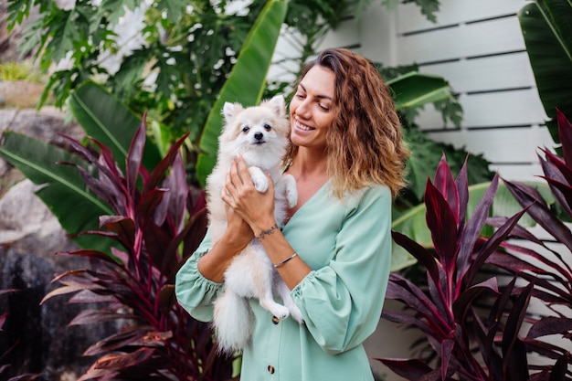 Retrato ao ar livre de uma mulher bronzeada europeia cacheada segurando um feliz spitz de cachorro de estimação