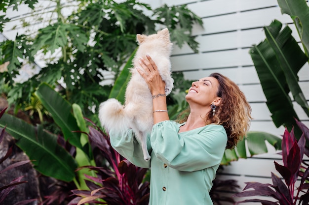 Retrato ao ar livre de uma mulher bronzeada europeia cacheada segurando um feliz spitz de cachorro de estimação