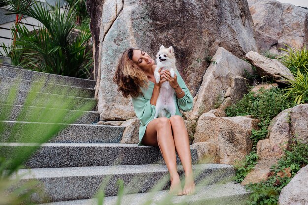 Retrato ao ar livre de uma mulher bronzeada europeia cacheada segurando um feliz spitz de cachorro de estimação