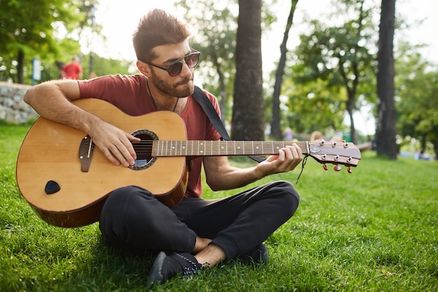 Retrato ao ar livre do cara bonito hipster sentado na grama do parque tocando violão