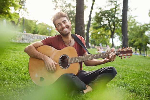 Retrato ao ar livre do cara bonito hipster sentado na grama do parque tocando violão