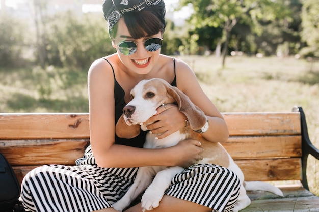 Retrato ao ar livre do close-up da fascinante garota da risada segurando o cachorro beagle enquanto está sentado no banco. jovem satisfeita de óculos escuros brincando com o cachorro no parque