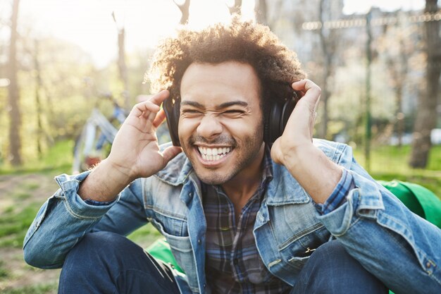 Retrato ao ar livre do close-up do homem africano bonito com cabelo afro, segurando as mãos em fones de ouvido enquanto ouve música e está animado, sentado no parque.