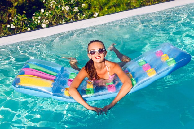 Retrato ao ar livre do estilo de vida de uma jovem deslumbrante se divertindo na piscina infinita com uma vista deslumbrante sobre a ilha tropical, usando biquíni brilhante e óculos escuros, nadando no colchão de ar.