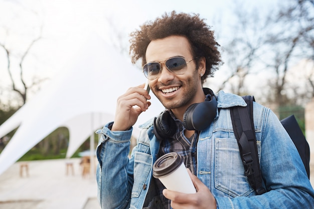 Retrato ao ar livre do homem de pele escura atraente elegante com penteado afro, usando fones de ouvido no pescoço, falando no smartphone e bebendo café enquanto passeava na cidade com a mochila.