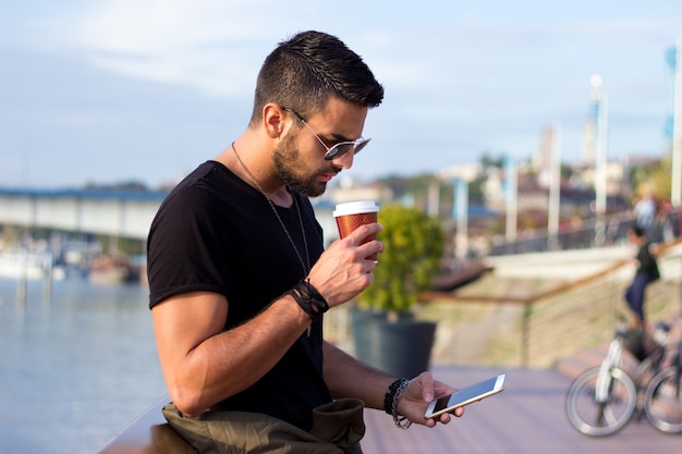 Retrato ao ar livre do homem novo moderno com o telefone móvel no café da terra arrendada da rua.