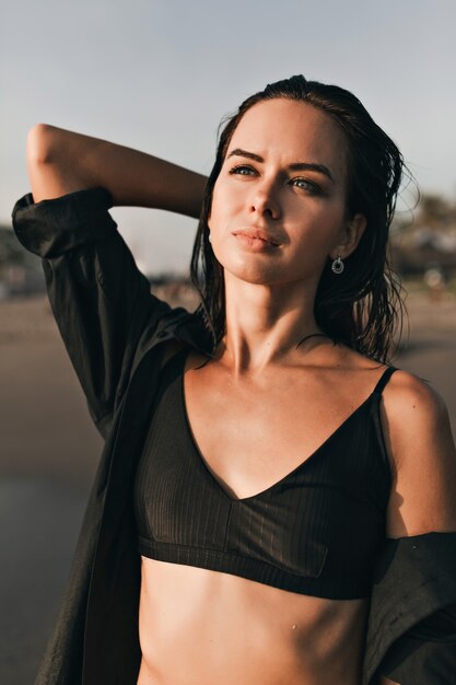 Retrato ao ar livre na moda de uma mulher bonita e atraente com cabelo escuro e olhos azuis, olhando para o oceano à luz do sol