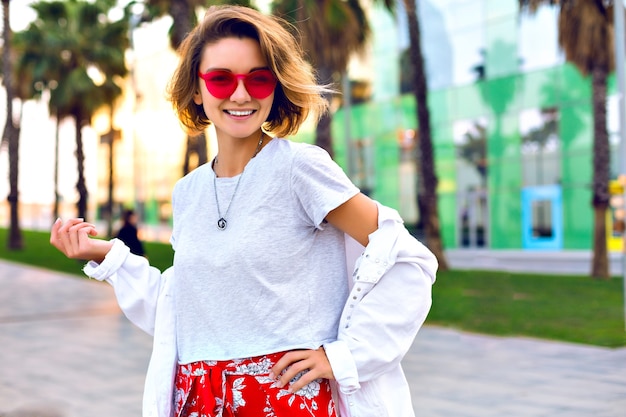 Retrato brilhante de moda ao ar livre de verão de mulher sorridente na moda elegante, vestindo roupa elegante de hipster, jaqueta jeans branca e óculos de sol de néon, palmas ao redor, clima de viagem feliz.