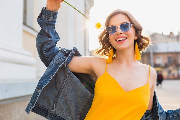 Retrato brilhante de uma linda mulher segurando uma flor, vestido amarelo, jaqueta jeans, estilo moderno, tendência da moda de verão, sorriso, óculos de sol da moda