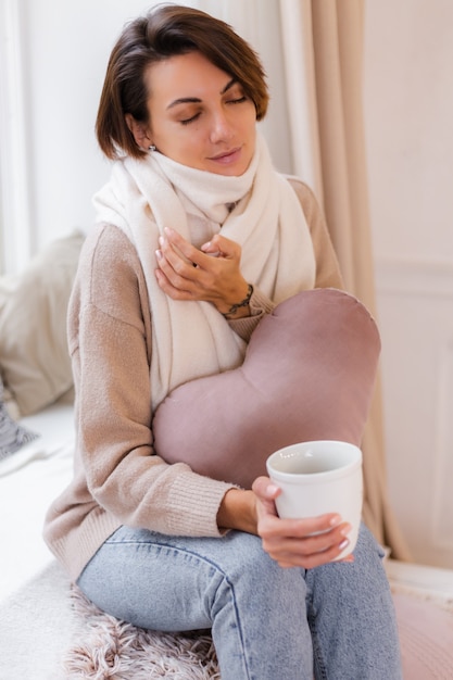 Retrato caloroso de mulher sentada no parapeito da janela com uma xícara de chá quente, café, suéter e lenço branco