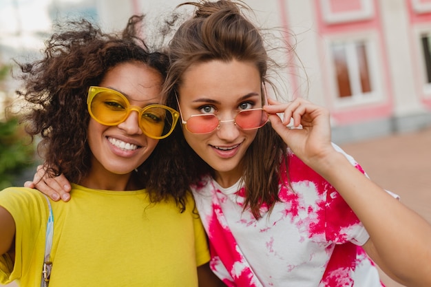 Retrato colorido de felizes amigas jovens sorrindo sentadas na rua tirando uma foto de selfie no celular, mulheres se divertindo juntas