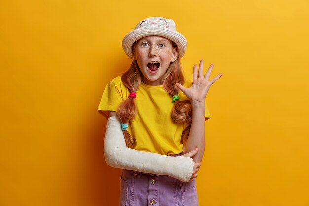 Retrato da alegre garota ruiva acena a palma da mão em um gesto de olá, diz oi para os pais, estando de bom humor, usa roupa de verão, elenco no braço quebrado após cair durante patins, isolado na parede amarela