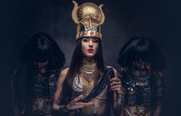 Retrato da altiva rainha egípcia em uma antiga fantasia de faraó com duas concubinas. Isolado em um fundo escuro.