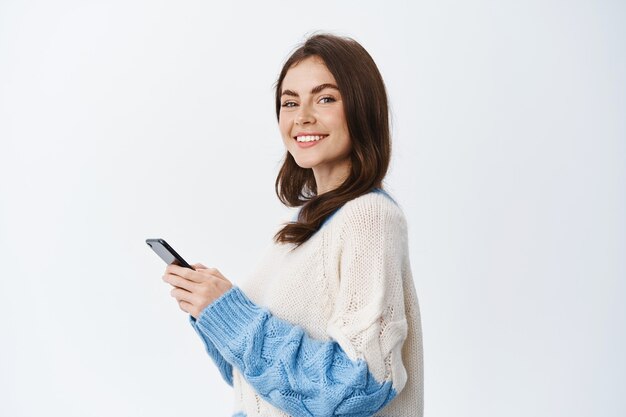 Retrato da bela modelo feminina usando o aplicativo de smartphone, em pé em seu perfil e virar o rosto com um sorriso na frente, enviando mensagem no chat de mídia social, parede branca