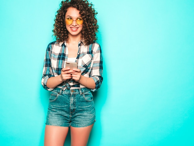 Retrato da bela modelo sorridente com penteado afro cachos, vestido com roupas de verão. Garota despreocupada posando perto de parede azul. Mulher usa seu telefone celular e digitando sms.