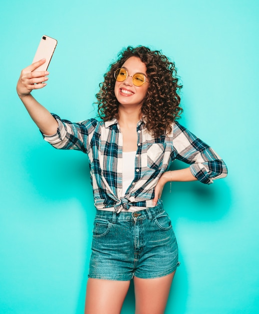 Retrato da bela modelo sorridente com penteados afro cachos, vestido com roupas de hipster de verão. Garota despreocupada sexy, posando no estúdio em fundo cinza. Mulher engraçada na moda tira foto de selfie