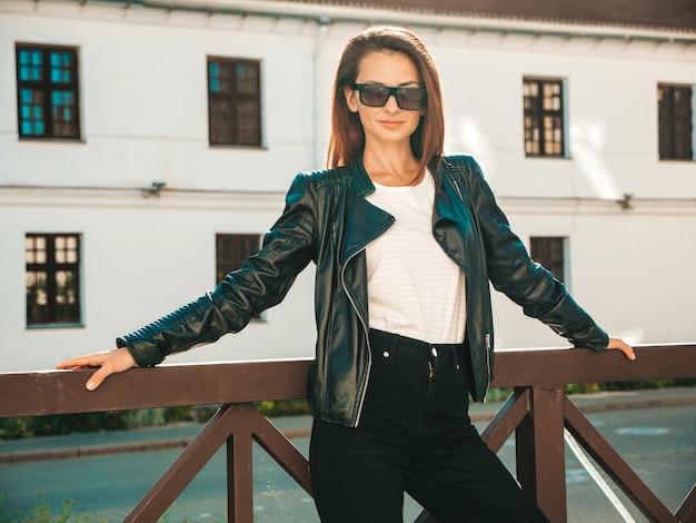 Retrato da bela modelo sorridente. mulher vestida com calça jeans e jaqueta de couro preto hipster de verão. mulher na moda posando na rua