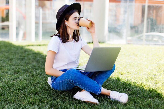 Retrato da bela mulher sorridente beber café sentado na grama verde no parque com as pernas cruzadas durante o dia de verão enquanto estiver usando o laptop