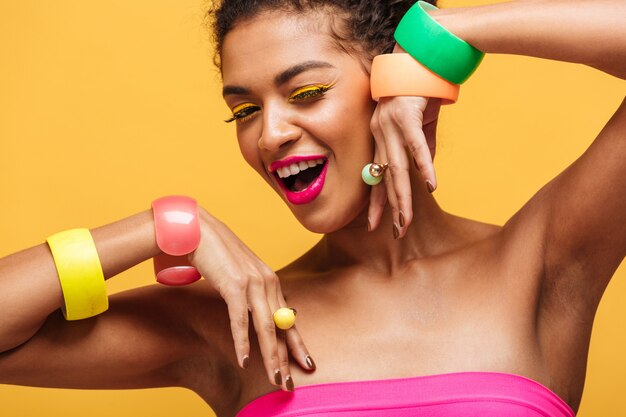 Retrato da beleza da mulher maravilhosa de raça mista com maquiagem moda e jóias nas mãos posando isolado, ao longo da parede amarela