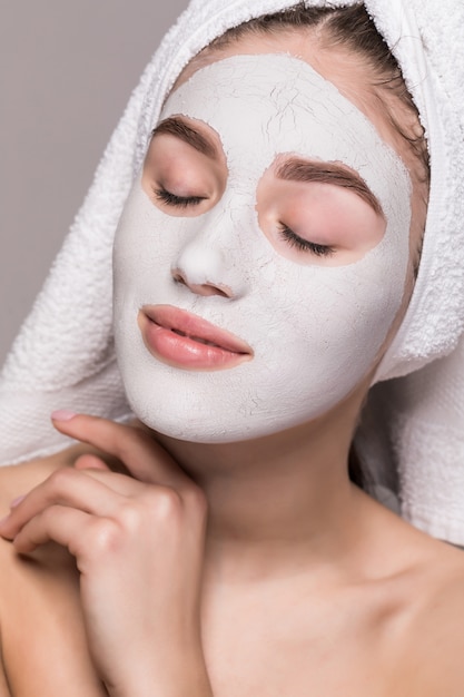 Retrato da beleza da mulher na toalha na cabeça com máscara de nutrição branca ou nata na cara, parede branca isolada. Skincare limpeza eco orgânico cosméticos spa relaxar conceito