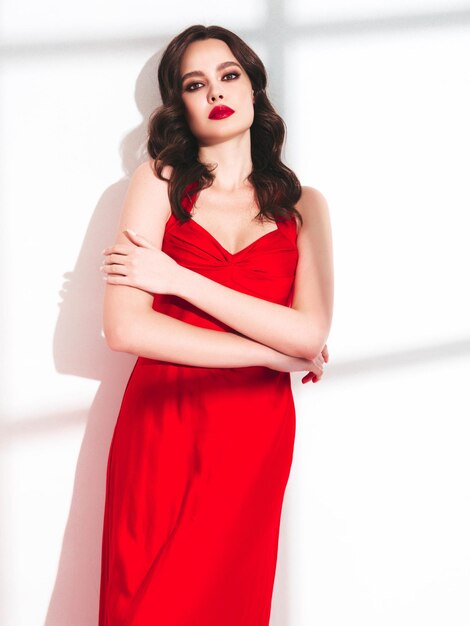 Retrato da beleza de uma jovem morena com maquiagem de noite e pele limpa perfeita Modelo sexy com cabelos cacheados posando no estúdio com lábios naturais brilhantes vermelhos em vestido vermelho