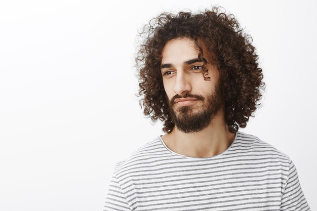 Retrato da cintura para cima de um lindo estilista masculino sonhador com corte de cabelo encaracolado e uma elegante camiseta listrada, olhando para longe e sorrindo sensualmente