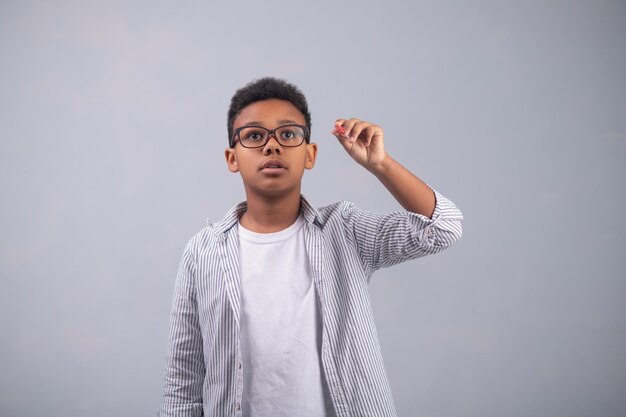 Retrato da cintura para cima de um menino concentrado em uma camisa listrada e óculos fazendo um esboço