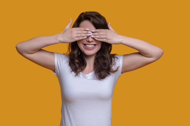 Retrato da cintura para cima de uma mulher caucasiana de cabelos escuros sorridente, cobrindo os olhos com as duas mãos