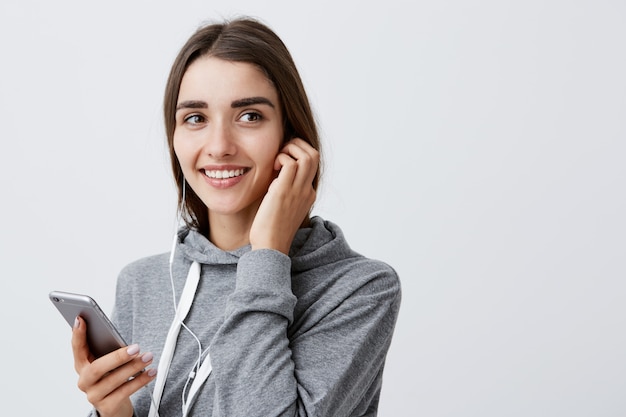 Retrato da encantadora aluna caucasiana morena linda com penteado longo com capuz cinza elegante, sorrindo com dentes, segurando o smartphone na mão, ouvindo música favorita em fones de ouvido