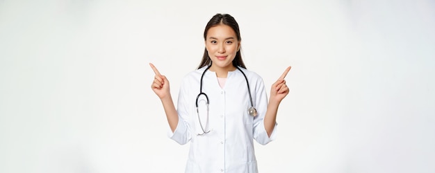 Retrato da médica asiática jovem profissional de saúde em roupão médico apontando para o lado mostrando tw