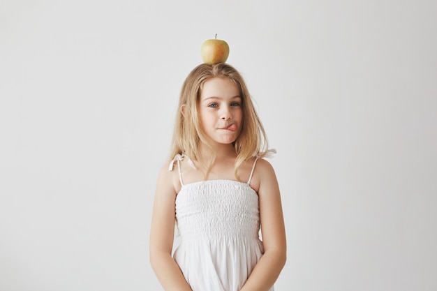 Retrato da menina alegre engraçada com cabelos loiros no vestido branco, segurando a maçã na cabeça, mostrando a língua, fazendo expressões tolas.