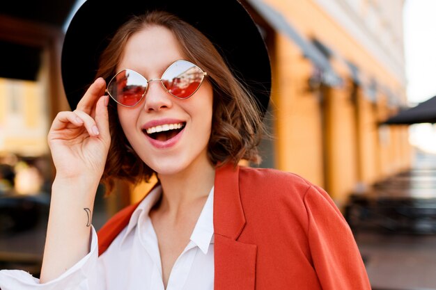 Retrato da menina européia de sorriso em óculos de sol alaranjados bonitos, jaqueta e chapéu preto. Moda outono. Café de rua.