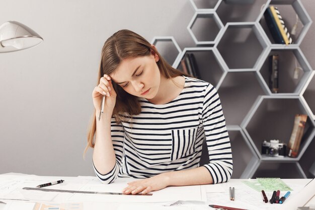 Retrato da menina freelance européia de cabelo escuro nova considerável no desenho da camisa listrada, olhando com expressão séria e concentrada da cara nos papéis, olhando através de seu trabalho.