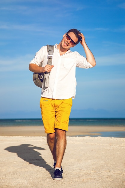 Retrato da moda ao ar livre de homem bonito em roupa casual da moda brilhante caminhando em uma praia tropical