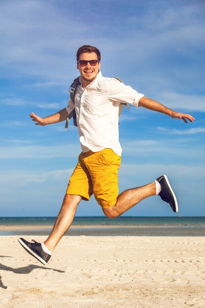 Retrato da moda ao ar livre de homem bonito em roupa casual da moda brilhante caminhando em uma praia tropical