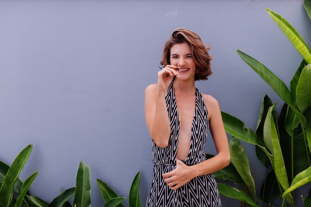 Retrato da moda da jovem modelo elegante caucasiana com macacão longo clássico posando na parede e folhas tropicais. Luz natural ao ar livre