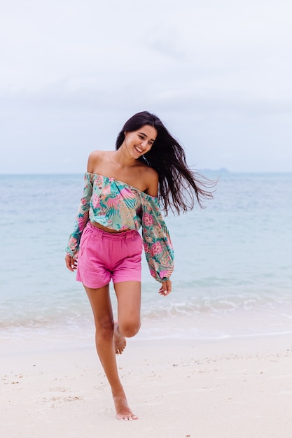 Retrato da moda da mulher elegante em top de manga comprida de impressão colorida e shorts rosa na praia, fundo tropical.