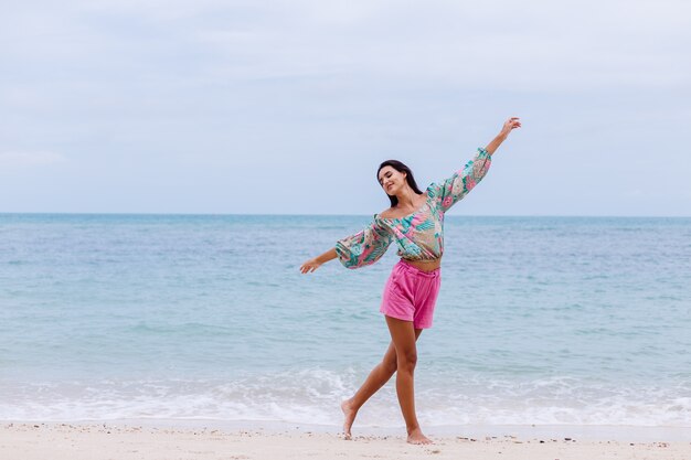 Retrato da moda da mulher elegante em top de manga comprida de impressão colorida e shorts rosa na praia, fundo tropical.