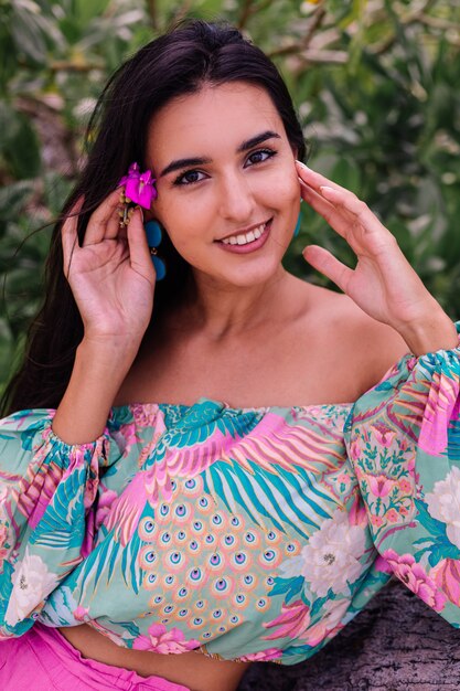 Retrato da moda da mulher elegante em top de manga comprida de impressão colorida e shorts rosa na praia, fundo tropical.