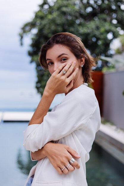 Retrato da moda da mulher em uma vila de luxo tropical usando um blazer branco elegante e joias sobre folhas tropicais