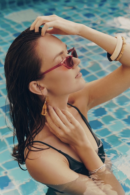 Retrato da moda da mulher graciosa sedutora em elegantes brincos amarelos com corpo perfeito posando na piscina durante as férias no resort de luxo.