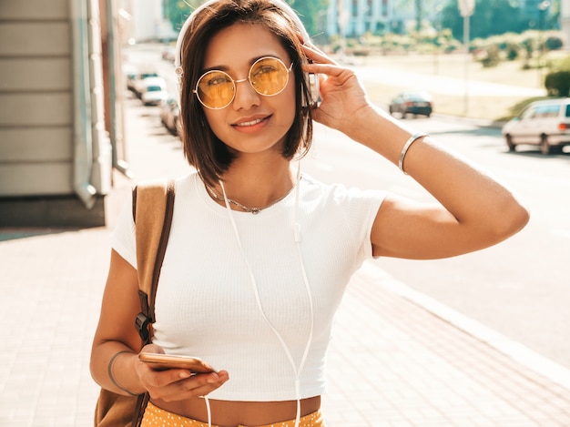 Retrato da moda da mulher jovem hippie elegante andando na rua. Garota vestindo roupas da moda bonito. Modelo sorridente desfrutar de seus fins de semana, viajar com mochila. Mulher ouvindo música através de fones de ouvido