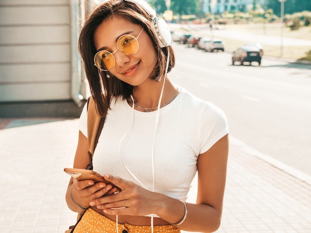 Retrato da moda da mulher jovem hippie elegante andando na rua. Garota vestindo roupas da moda bonito. Modelo sorridente desfrutar de seus fins de semana, viajar com mochila. Mulher ouvindo música através de fones de ouvido