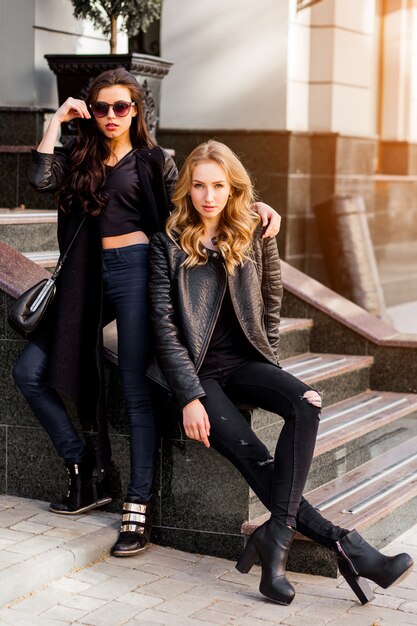 Retrato da moda de duas mulheres bonitas elegantes, posando na rua em um dia ensolarado. Vestindo roupa urbana da moda, jaqueta de couro e botas de salto alto. Jovens amigos esperando na escada ao ar livre.