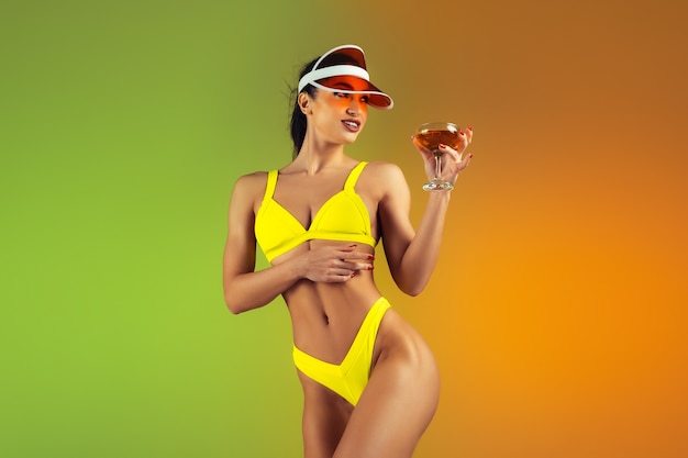 Retrato da moda de jovem em forma e mulher esportiva com coquetel em um elegante maiô de luxo amarelo na parede gradiente, corpo perfeito pronto para o verão