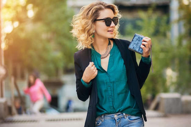 Retrato da moda jovem elegante andando na rua com uma jaqueta preta, blusa verde, acessórios elegantes, segurando uma bolsa pequena, usando óculos escuros, estilo de moda de rua de verão