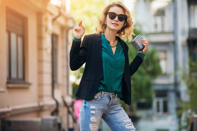 Retrato da moda jovem elegante andando na rua com uma jaqueta preta, blusa verde, acessórios elegantes, segurando uma bolsa pequena, usando óculos escuros, estilo de moda de rua de verão