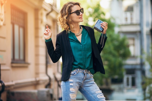 Retrato da moda jovem elegante andando na rua com uma jaqueta preta, blusa verde, acessórios elegantes, segurando uma bolsa pequena, usando óculos escuros, estilo de moda de rua de verão