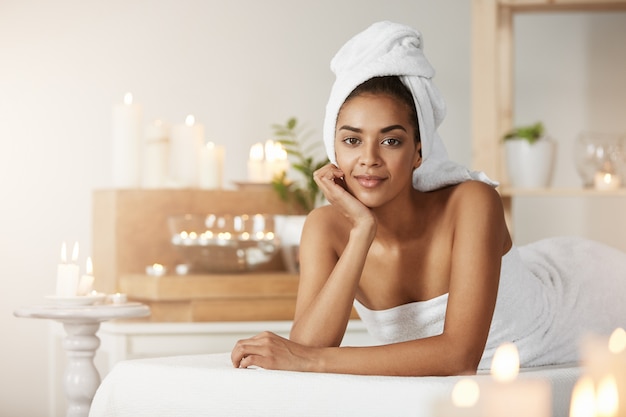 Retrato da mulher africana bonita com a toalha na cabeça que sorri descansando no salão spa.